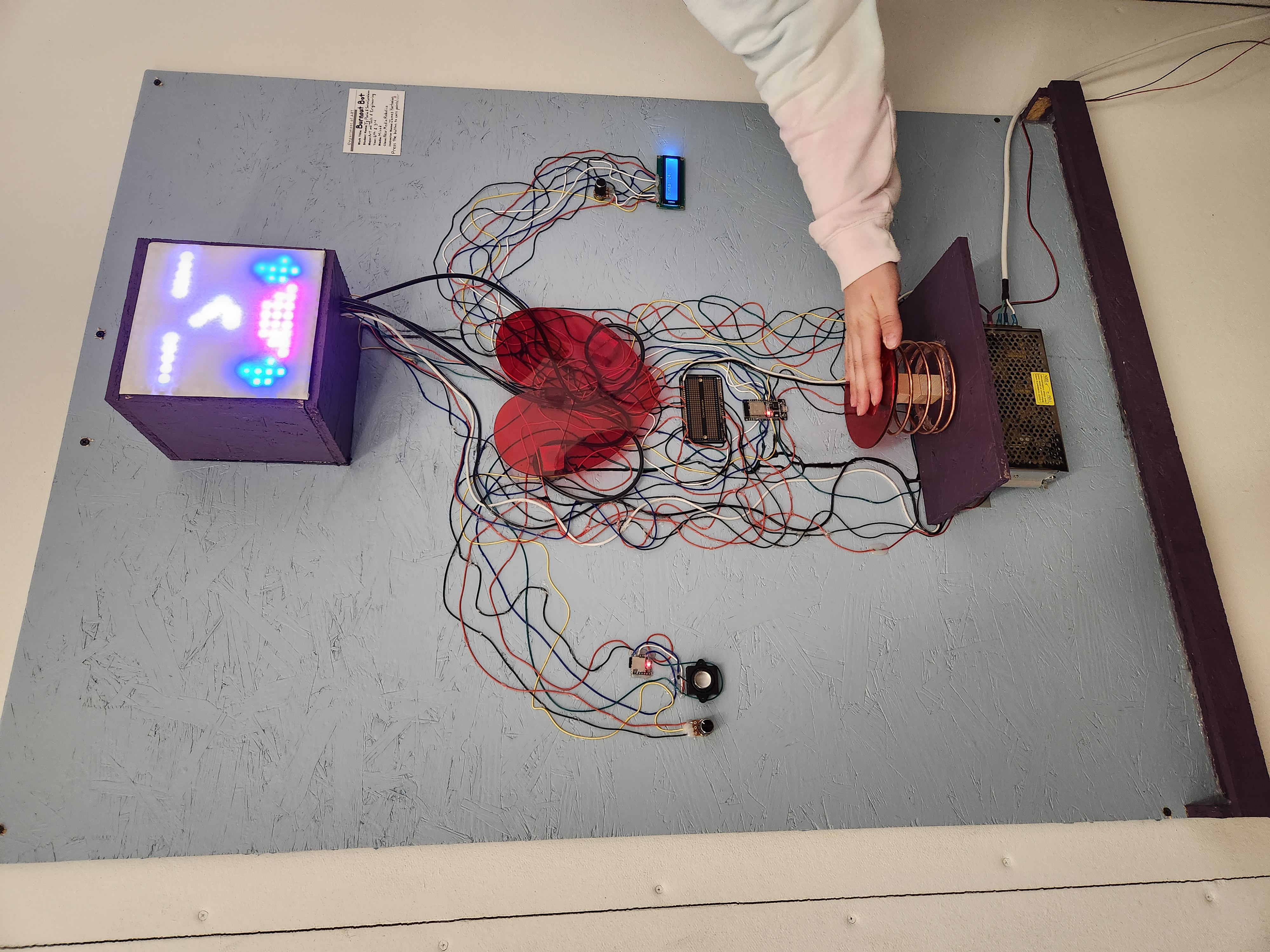 Photo with a hand pressing a large button on the Burnout Bot, which has been installed on a wall. The Burnout Bot is composed of wire art connecting several electronic components, a tubing system carrying oil, and other materials used for aesthetic or construction purposes. In this photo, the Burnout Bot's face screen has a simple crying face animation.